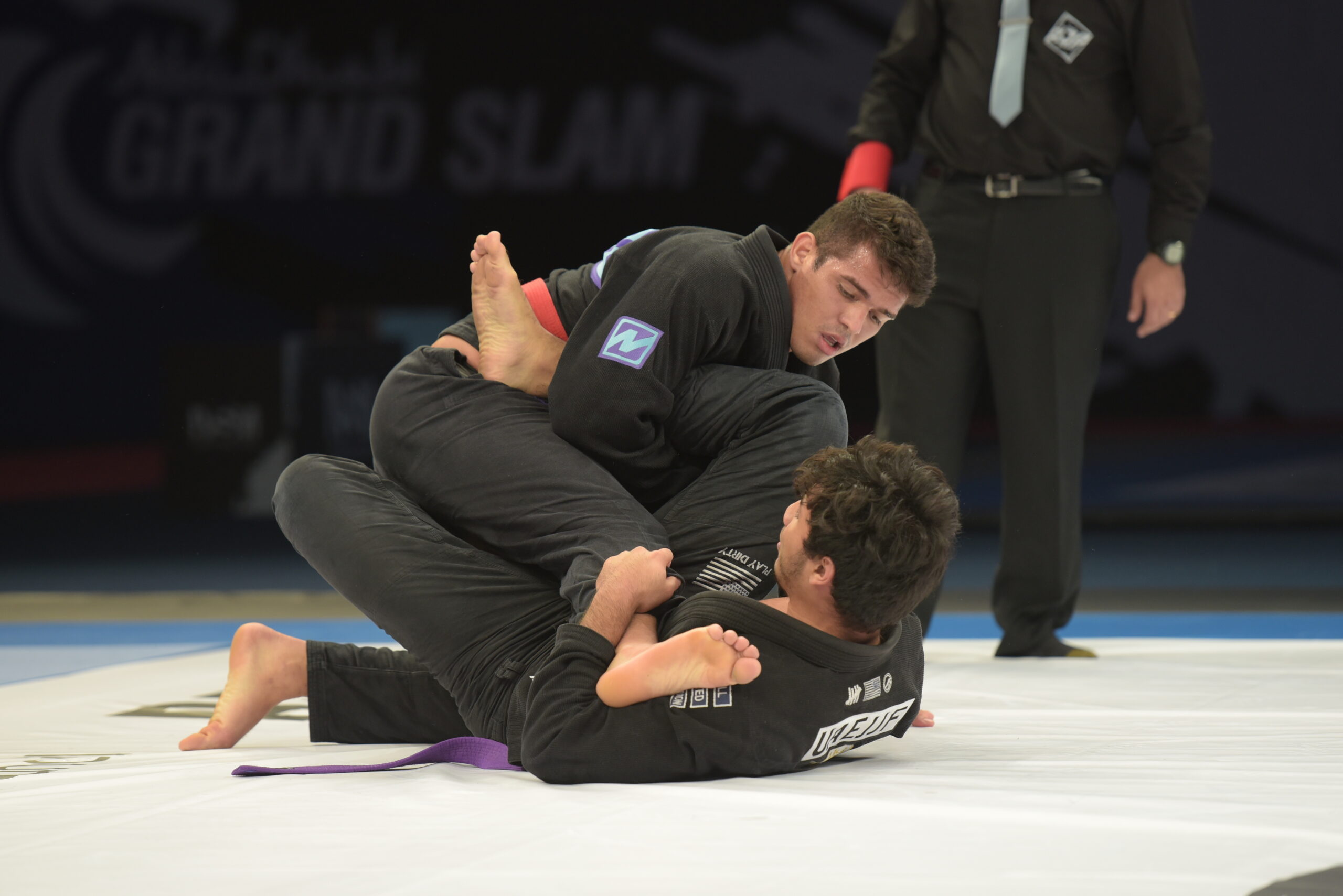 Dois atletas de quimono preto disputam uma posição no tatame, acompanhados por um árbitro.